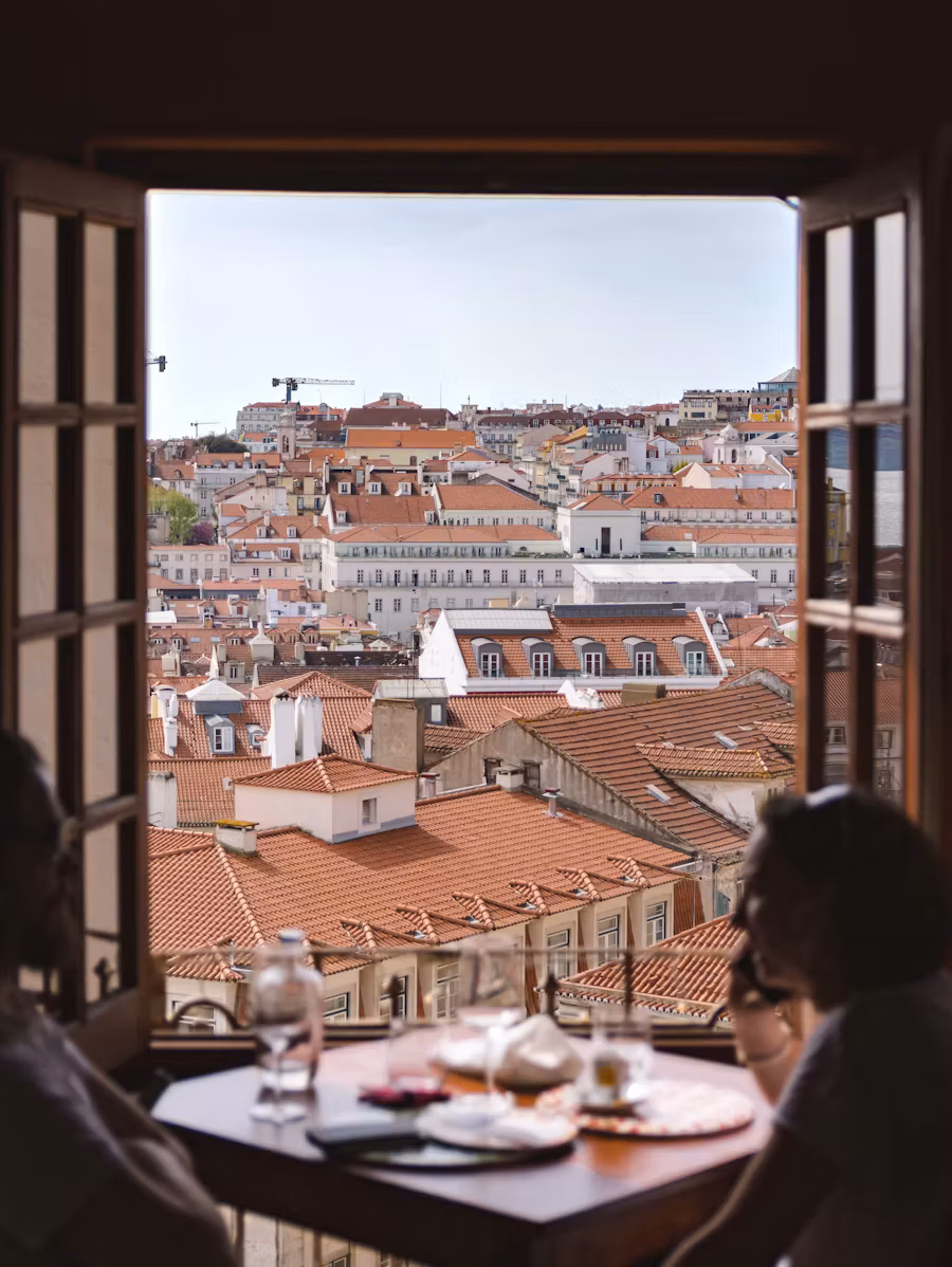 Open window over Lisbon’s terracotta rooftops, two people seated at a table in the foreground